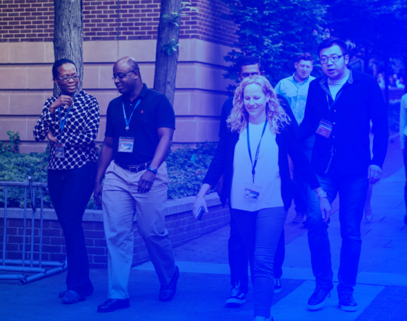 A group of people wearing lanyards, walking along a brick building, some smiling and engaged in conversation. The scene has a blue tint.