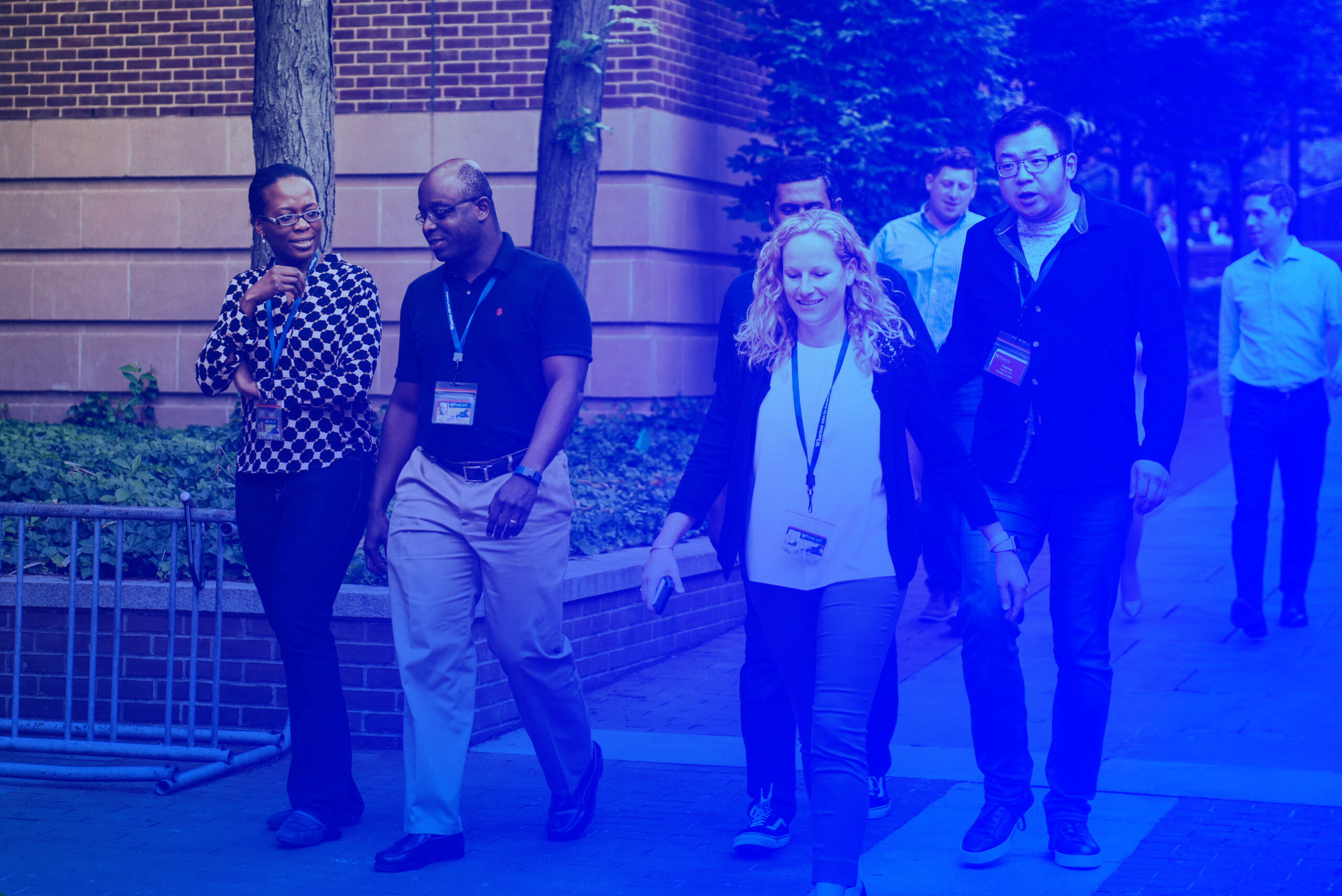 A group of people wearing lanyards, walking along a brick building, some smiling and engaged in conversation. The scene has a blue tint.