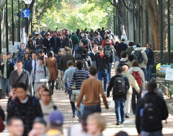 A busy outdoor walkway with a large group of people, many carrying backpacks, surrounded by trees and lamp posts.